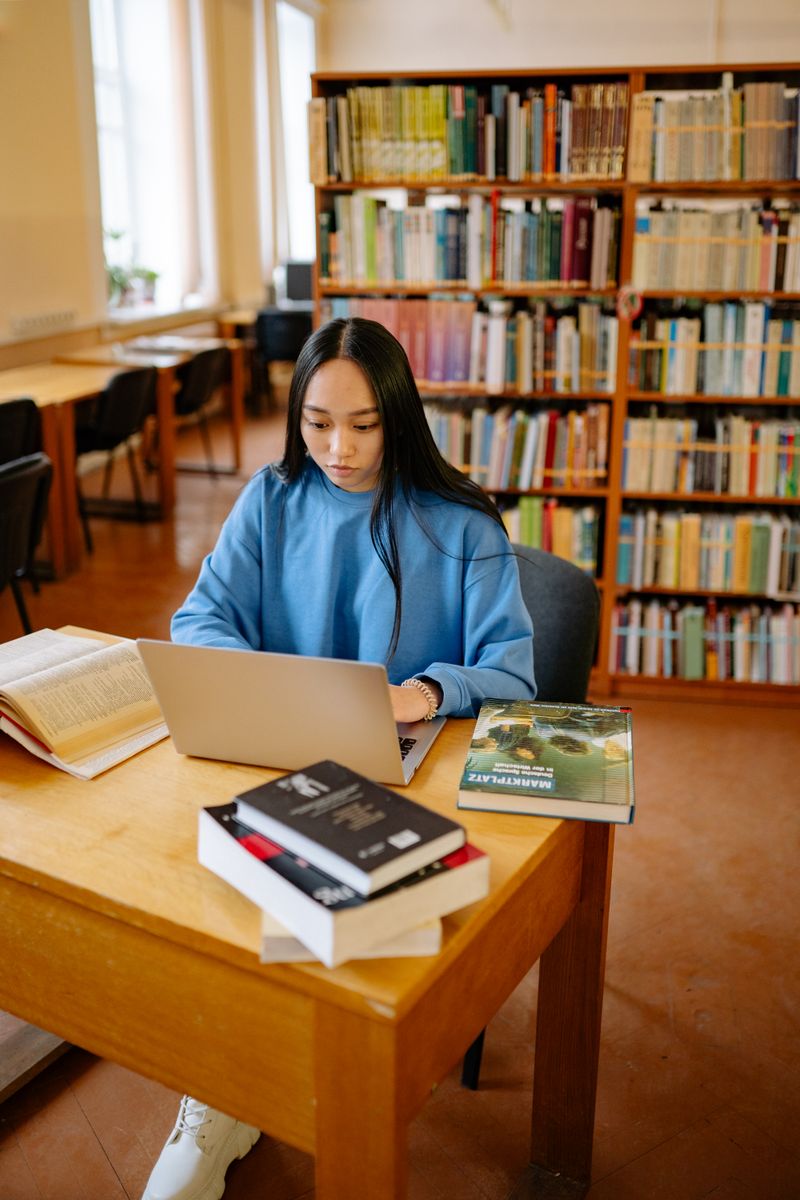 Holiday Camp 3 Young woman in library using laptop surrounded by books, focused on studying.